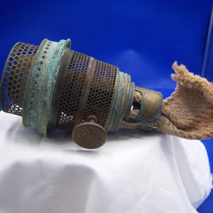 May include: An antique oil lamp burner with a perforated metal body and a wick. The burner has a brass finish with a green patina, and a small knob. The wick is made of a tan, woven material. The lamp is set against a blue background.