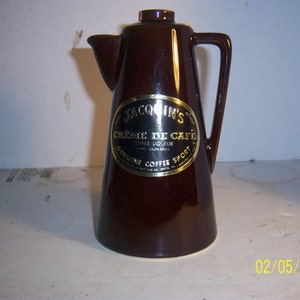 May include: A brown ceramic coffee pot with a handle and spout. The pot has a gold label with the text "Jacquin's Crème de Café Coffee Liqueur". The pot is a vintage style and is likely used for serving coffee or other beverages.