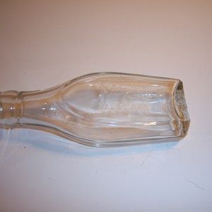 May include: A clear glass bottle with a textured surface. The bottle is empty and has a slightly rounded shape.