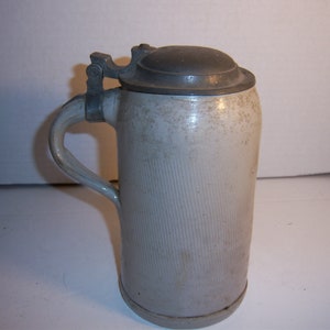 May include: A white ceramic beer stein with a pewter lid and handle. The stein has a textured surface and is in good condition.