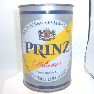 May include: A vintage silver beer can with blue and yellow accents. The can features the word "PRINZ" in large blue letters, with "Pilsener" in red script below. The can also has a crest and text in German.
