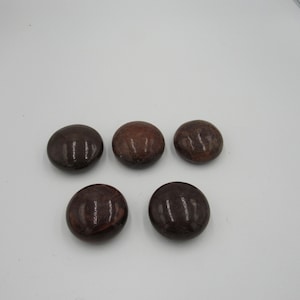 May include: Five polished, round stones in varying shades of brown and burgundy are arranged on a white surface. The stones have a smooth, glossy finish, and their colors range from dark chocolate to reddish-brown.