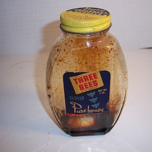 May include: A glass jar of Three Bees Clover Pure Honey. The jar is brown and has a yellow lid. The label is blue with white text and a picture of three bees.