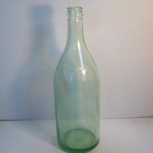 May include: A clear glass bottle with a green tint. The bottle has a rounded bottom and a long neck. The bottle is empty and has no label.