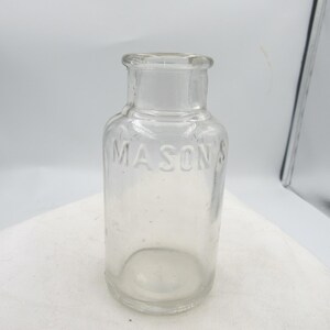 May include: A clear glass bottle with the embossed word "MASON'S" on the front. The bottle has a rounded body, a narrow neck, and a small opening. The bottle is sitting on a white surface.