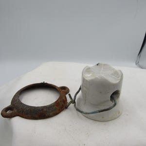 May include: A vintage electrical insulator with a white ceramic body and a rusty metal ring. The insulator has a cylindrical shape with a rounded top and a hole for wiring. The metal ring is circular with two small loops.