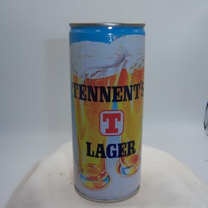 May include: A cylindrical Tennent's Lager beer can. The can features a blue top, with the brand name and logo in the center. The background shows a depiction of beer glasses with foam. The word "LAGER" is printed below the logo.