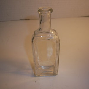 May include: A clear glass bottle with a square body and a narrow neck. The bottle has a textured surface and is empty.