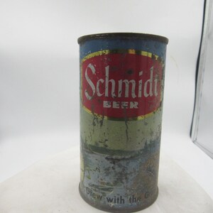 May include: A vintage Schmidt Beer metal can featuring a red oval logo with white text on a blue and light gray background. The cylindrical container shows signs of wear, rust, and surface oxidation, with the text Brew with the Great Outdoors.