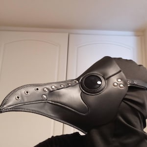 May include: A black plague doctor mask featuring a long, pointed beak, circular eye lenses with dark glass, and silver-toned metal rivets along the top and side. The mask is constructed with stitched seams and includes an adjustable strap.