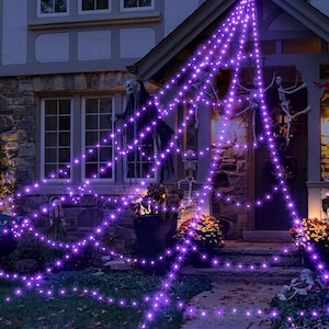 Puede incluir: Una gran decoración de telaraña triangular iluminada por luces LED de color púrpura brillante, colgada del techo de un porche. La red presenta varias figuras de esqueletos de plástico unidas a los hilos, creando una exhibición festiva.