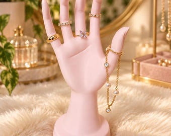Op de afbeelding: Een lichtroze etalagehand als displaystandaard met vier goudkleurige ringen en een goudkleurige kettingarmband. De ringen omvatten een gladde band, een met steentjes bezette band, een vlindervormige ring en een dunne band. De armband is voorzien van kleine parels en heldere steentjes.