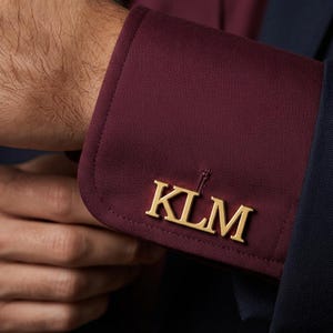 May include: A gold-toned metal cufflink featuring the letters KLM in a serif font, attached to the burgundy fabric cuff of a long-sleeved shirt. The cufflink is positioned horizontally across the center of the visible fabric cuff.