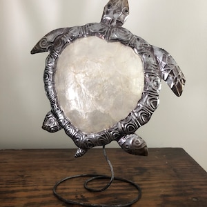 May include: A silver metal turtle sculpture with a white mother of pearl shell on its back. The turtle is standing on a black metal stand.