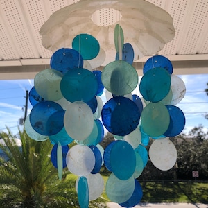 May include: A white seashell wind chime with blue and green iridescent seashells hanging from it. The wind chime is hanging from a white ceiling.