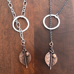 May include: Two necklaces with a leaf pendant. One necklace has a silver chain and a white circle, the other has a black chain and a silver circle. The leaf pendants are copper-colored with a textured design and a small copper-colored ball.