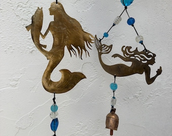 May include: Two brass mermaid wind chimes with blue and clear beads. One mermaid holds a fish, the other is swimming. Each chime has a small bell at the bottom. A decorative nautical theme.