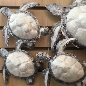 May include: Several decorative sea turtle figurines with silver-colored trim and textured white shells are displayed. The turtles vary in size and are mounted on a light brown wooden surface. The turtles' flippers and heads are detailed with intricate patterns.