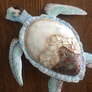 May include: A blue and white ceramic turtle sculpture with a detailed shell made of iridescent mosaic tiles. The turtle is standing on a white pedestal.