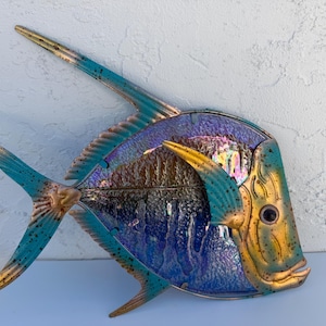 May include: A metal wall art piece in the shape of a fish. The fish has a teal, gold, and blue color scheme. The fins and tail are teal, while the body has a mix of gold, blue, and iridescent colors. The fish has a large eye.