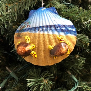 May include: A seashell Christmas ornament painted to look like a beach scene with two sea turtles on the sand. The seashell is blue and white with a blue wave at the top. The turtles are brown with yellow spots.