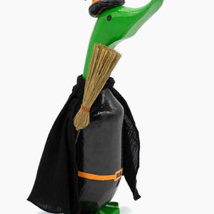 May include: A green duck figurine wearing a black witch hat with an orange band, a black cape, and an orange belt. The duck holds a small tan straw broom against its chest and stands on green feet against a plain white background.