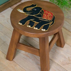 May include: A small round wooden stool with four legs and a circular seat. The top of the seat features a hand-painted black elephant with white tusks and a red decorative blanket with gold-coloured swirling patterns outlined in gold paint.