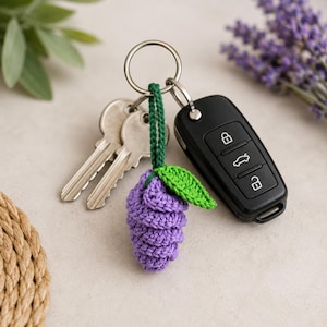 May include: A metal keyring holding two silver-coloured keys, a black car key fob with three buttons, and a small, handmade purple crocheted grape cluster charm featuring a single green crocheted leaf attached by a dark green yarn loop.