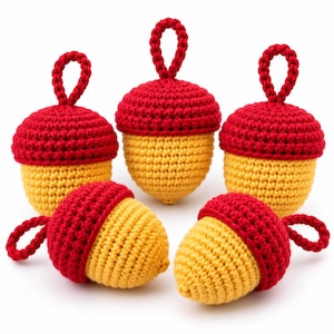 May include: A set of five crocheted acorn ornaments, each featuring a bright yellow base and a textured red cap. Three acorns are positioned upright with a small red loop at the top, while two acorns are lying on their sides with a red loop attached.
