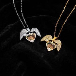 Puede incluir: Dos collares con relicario en forma de corazón con alas de ángel, uno en metal plateado y otro en dorado. Cada relicario contiene una pequeña fotografía de una pareja y está adornado con cristales transparentes en las alas y el marco.