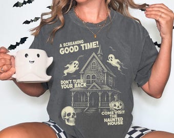 May include: Dark gray t-shirt with a Halloween-themed graphic. The design features a haunted house, ghosts, a skull, and a pumpkin. Text on the shirt reads "A Screaming Good Time!" and "Come Visit The Haunted House". A white ghost-shaped mug is held in the image.