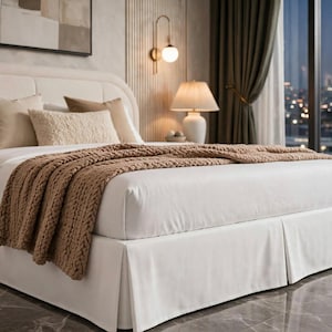 May include: A white bed skirt with a tailored, pleated design that covers the base of the bed. The fabric is smooth and opaque, extending from the mattress down to the floor, creating a clean, finished look for the bed frame and foundation.