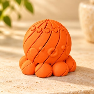 Peut inclure: Une figurine de pieuvre articulée, sphérique et orange vif, présentant un design segmenté en spirale avec une finition mate lisse. L'objet possède une tête arrondie et plusieurs petits tentacules arrondis à la base, tous dotés de joints articulés visibles.