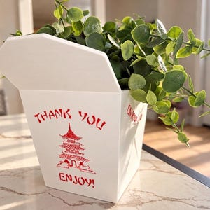 May include: A white takeout container with a red pagoda graphic and the red text "THANK YOU" and "ENJOY!" printed on the front. The container is filled with a green leafy plant with small round leaves, extending out from the open top.