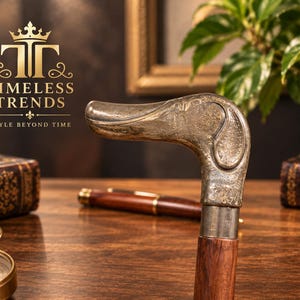 Può includere: Un bastone da passeggio con un'impugnatura metallica a forma di testa di anatra con dettagli incisi complessi. L'impugnatura è fissata a un bastone in legno liscio marrone scuro. Il testo TIMELESS TRENDS STYLE BEYOND TIME è visibile in alto a sinistra.