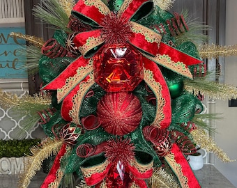 May include: A festive Christmas wreath with a green mesh base, adorned with red velvet bows, gold accents, and red and green ornaments. The wreath features a large, faceted red jewel at its center, creating a striking holiday decoration.