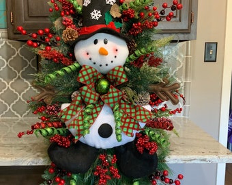 May include: A festive snowman decoration with a black top hat adorned with snowflakes and a red band. The snowman features a white body, black coal eyes and mouth, and an orange carrot nose. It is decorated with red berries, green ribbon, and pinecones.