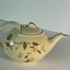 May include: A cream-colored ceramic teapot featuring a rounded body, a curved handle, and a matching lid with a gold-trimmed finial. The exterior is decorated with a botanical pattern of stylized leaves and flowers in shades of brown, yellow, and tan.