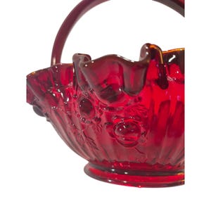 May include: A deep red glass basket featuring a ruffled, scalloped rim and a curved handle. The exterior surface of the bowl is decorated with a moulded floral pattern of roses and leaves, set against a vertical ribbed texture on a circular base.