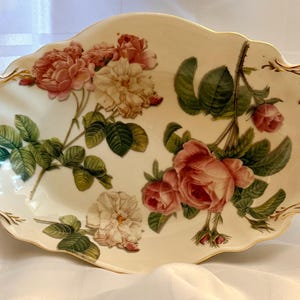 May include: An oval porcelain serving dish with a cream-coloured base, featuring a detailed floral pattern of pink and white roses with green leaves. The dish has ornate, openwork handles on both sides accented with gold trim along the edges.