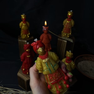 May include: A yellow and red wax candle shaped like a figure wearing a headwrap and traditional clothing with beaded accents. The candle features intricate molded details, including a textured skirt and bodice, with a white wick at the top.