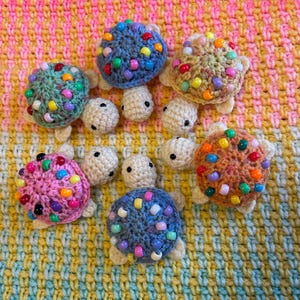 May include: Six small crocheted turtles with cream-coloured heads and limbs. Each turtle features a uniquely coloured shell, including blue, green, pink, tan, and orange, all decorated with various multi-coloured plastic beads attached to the yarn.