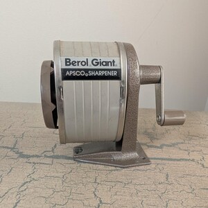 May include: A vintage Berol Giant Apsco pencil sharpener featuring a textured brown metal base and crank handle, paired with a light gray cylindrical body. The front label clearly displays the text Berol Giant and APSCO SHARPENER in black.