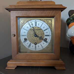 May include: A wooden mantel clock with a square frame and a brass handle on top. The clock face features a gold-toned center with intricate floral patterns, black Roman numerals, and the text Marshall Field & Company printed in the center.