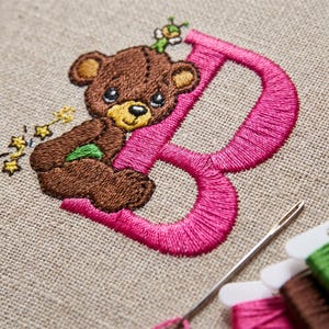May include: An embroidery design on beige fabric featuring a large, bright pink letter B. A brown teddy bear sits against the letter, accompanied by a small green insect on its head and yellow star-shaped accents. A metal needle lies nearby.