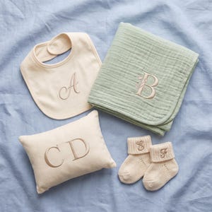 May include: A cream-colored baby bib with an embroidered letter A, a sage green muslin blanket with an embroidered letter B and floral detail, a cream rectangular pillow with embroidered letters CD, and two cream knit baby booties with letters E and F.