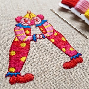 May include: An embroidery design on beige fabric featuring two clowns with pink and yellow striped sleeves and red trousers with yellow polka dots. The clowns wear blue ruffled collars and yellow hats, joined at the hands. A silver needle lies nearby.