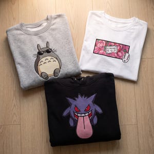Puede incluir: Una sudadera gris de cuello redondo con un personaje de dibujos animados con gafas de sol, una camiseta blanca con un gráfico floral rosa y un icono de máscara, y una sudadera negra con capucha con un personaje morado sacando la lengua.