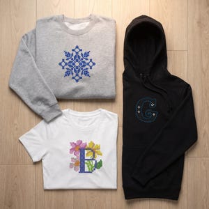 May include: A light gray crewneck sweatshirt with a blue cross-stitch snowflake, a white t-shirt featuring a blue letter B surrounded by pink and yellow flowers, and a black hooded sweatshirt with a blue cross-stitch letter G with white accents.
