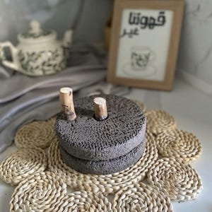 Peut inclure: Un moulin à main circulaire en pierre gris foncé poreuse, composé de deux disques de broyage texturés empilés, avec deux poignées cylindriques en bois clair dépassant de la surface supérieure du disque supérieur.